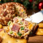 A close-up of a cheese knife spreading creamy, bacon-flecked cheese from a pecan-coated cheese ball onto a cracker.