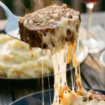 A close-up of a spatula lifting a thick slice of meatloaf, showing a long string of melted cheese and a juicy interior with mashed potatoes in the background.