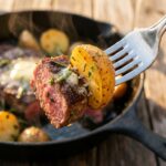 A close-up of a fork holding a juicy seared steak bite and a crispy potato coated in garlic butter, with a skillet and wine in the background.