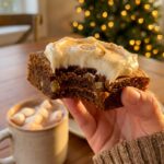 A close-up of a hand holding a frosted gingerbread bar with a bite taken out, showing the chewy texture, with a Christmas tree in the background.