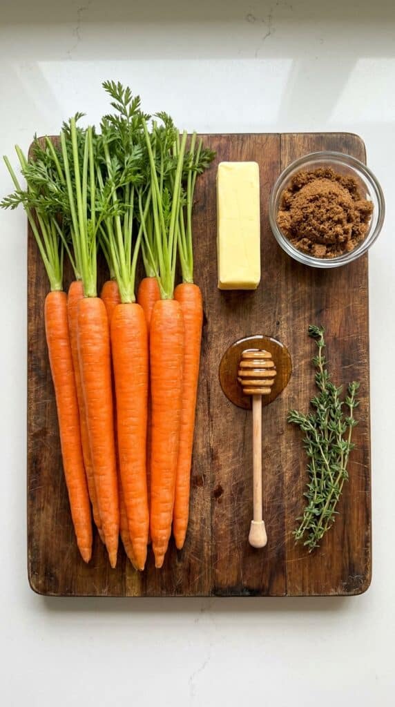 A flat lay showing whole carrots with green tops, butter, brown sugar, honey, and fresh thyme on a dark wooden board.