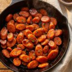 A close-up of a serving spoon lifting thick, glossy glazed carrots from a white bowl, with a sticky syrup dripping down.