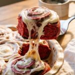 A close-up of a spatula lifting a gooey red velvet cinnamon roll from a pan, showing a stretch of melting white frosting and bright red dough.