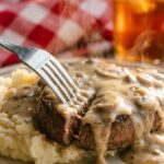 A close-up of a fork cutting through a tender ground beef steak covered in mushroom gravy, served over mashed potatoes.