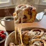 A close-up of a spatula lifting a scoop of gooey, hot cinnamon roll casserole with melting frosting dripping down.