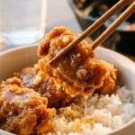 A close-up of wooden chopsticks holding a piece of glossy, sticky honey butter chicken over a bowl of white rice.