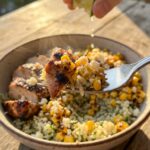 A close-up of a fork holding a bite of creamy street corn, chicken, and rice, with a hand squeezing a lime in the blurred background.