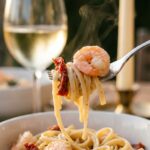 A close-up of a fork twirling creamy linguine with a seared shrimp and sun-dried tomato, with a glass of white wine in the background.
