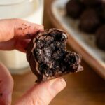 A close-up of a hand holding an Oreo truffle with a bite taken out, revealing a dense, dark, fudgy center, with a glass of milk in the background.