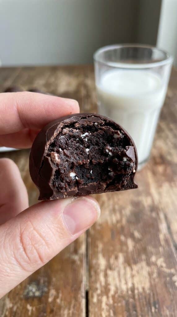A close-up of a hand holding an Oreo ball with a bite taken out, revealing a dense, dark fudgy center with a crisp chocolate shell.