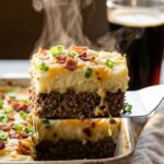 A close-up of a spatula lifting a square slice of meatloaf casserole, showing the distinct layers of beef and mashed potatoes.