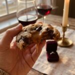 A close-up of a hand holding a half-eaten caramel-stuffed chocolate chip cookie, with a romantic candlelit dinner and a velvet ring box blurred in the background.