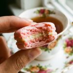 A close-up of a hand holding a pink macaron with a bite taken out, showing the chewy interior and buttercream filling, with a teacup in the background.