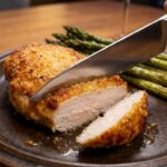 A close-up of a knife slicing into a juicy, parmesan-crusted chicken breast, revealing moist white meat inside.