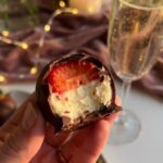 A close-up of a hand holding a chocolate-dipped strawberry with a bite taken out, revealing a white cheesecake center and juicy red berry.
