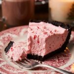 A close-up of a slice of pink peppermint pie with a chocolate crust on a festive plate, showing a fluffy texture with candy cane pieces.