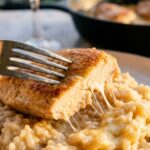 A close-up of a fork cutting into seared chicken over a bed of glossy, creamy parmesan rice on a plate.