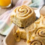 A close-up of a spatula lifting a gooey orange roll from a pan, showing a stretch of melted glaze and soft dough spirals.