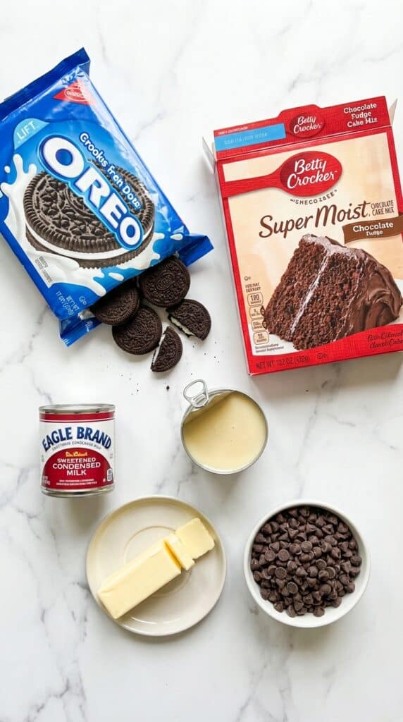 A flat lay showing whole Oreo cookies, a box of chocolate cake mix, sweetened condensed milk, sliced butter, and chocolate chips on a marble board.