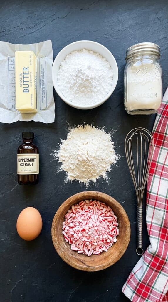 A flat lay showing butter, powdered sugar, cornstarch, peppermint extract, flour, and crushed candy canes on a dark slate board.