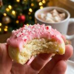 A close-up of a hand holding a peppermint meltaway cookie with a bite taken out, showing a crumbly, buttery interior, with hot chocolate and a Christmas tree in the background.