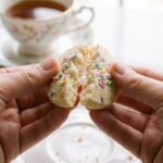 A close-up of a hand breaking a delicate shortbread cookie in half, showing a tender crumb, with a cup of tea in the background.