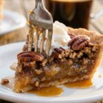 A close-up of a fork cutting into a slice of pecan pie topped with melting vanilla ice cream, showing the gooey amber center.