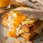 A close-up of a knife breaking a fried egg yolk on top of a savory puff pastry tart, with the bright orange yolk running over the cheese and ham.