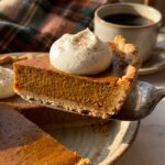 A close-up of a pie server lifting a thick, smooth slice of pumpkin pie topped with whipped cream, showing the flaky crust.