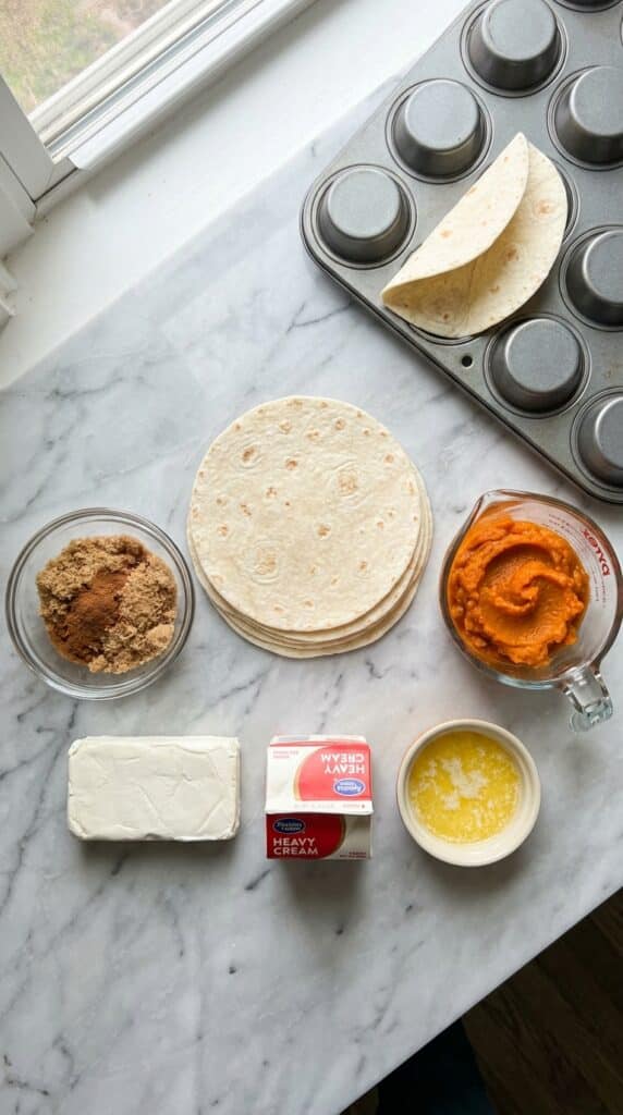 A flat lay showing mini tortillas, pumpkin puree, cream cheese, cinnamon sugar, and an upside-down muffin tin on a marble board.