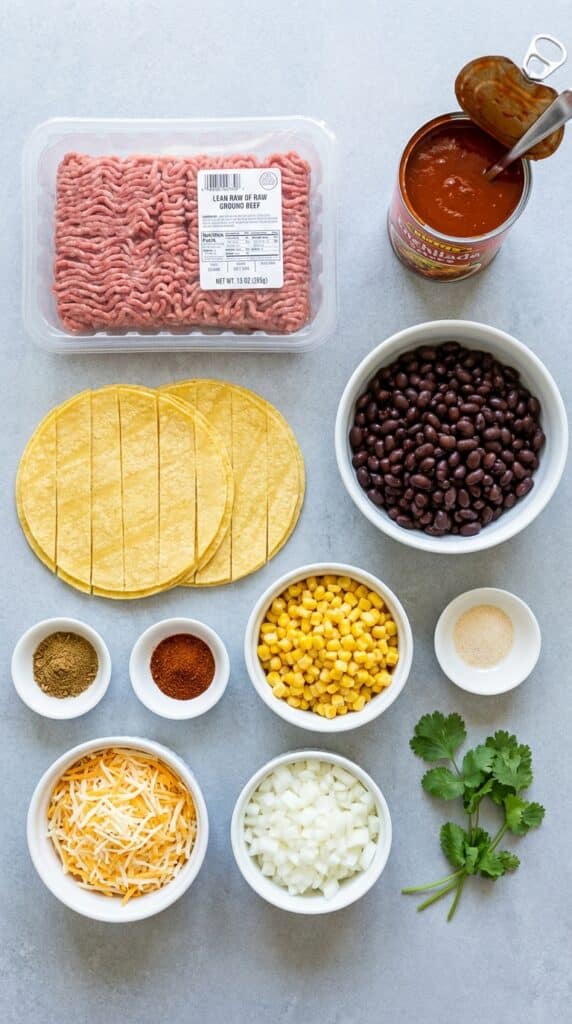 A flat lay showing raw ground beef, cut corn tortillas, enchilada sauce, black beans, corn, cheese, and diced onions on a slate board.