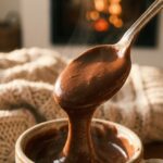 A close-up of a silver spoon holding a thick, glossy scoop of Italian hot chocolate, dripping slowly, with a fireplace in the background.