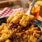 A close-up of a serving spoon lifting a large, cheesy scoop of tater tot and ground beef casserole, with a long cheese pull.