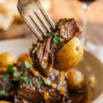 A close-up of a fork holding a tender bite of beef and a baby potato dripping with garlic butter sauce, with a bowl of food and red wine in the background.