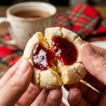 A close-up of hands breaking a jam-filled shortbread thumbprint cookie in half, showing the tender crumb and glossy red center.