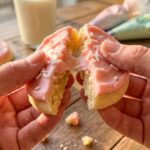 A close-up of hands breaking a thick, frosted sugar cookie in half, showing a very soft and tender interior crumb.
