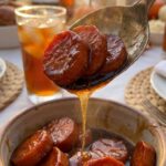 A close-up of a serving spoon lifting glazed sweet potatoes, showing a thick drip of caramelized brown sugar syrup falling from the spoon.