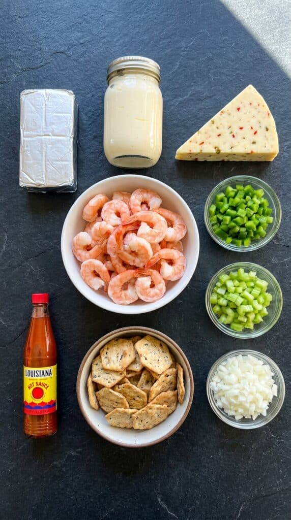 A flat lay showing cooked pink shrimp, cream cheese, hot sauce, pepper jack cheese, diced bell peppers, onions, celery, and crackers on a slate board.