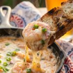 A close-up of a toasted baguette slice scooping up hot, creamy shrimp dip with melted cheese and green onions, with steam rising.