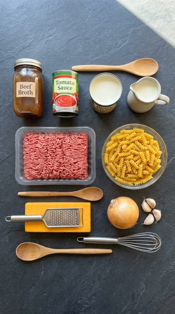 A flat lay showing raw ground beef, dry rotini pasta, beef broth, tomato sauce, cream, cheddar cheese, onion, and garlic on a dark slate board.
