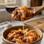 A close-up of a spoon lifting a bite of creamy ground beef and rotini pasta from a bowl, showing the thick sauce coating the noodles.