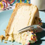 A close-up of a fork slicing into a dense, creamy piece of sugar cookie cheesecake on a blue plate, highlighting the smooth texture and thick cookie crust.