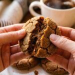 A close-up of hands breaking a soft molasses cookie in half, revealing a dense, chewy center, with a mug of tea in the background.