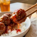 A close-up of chopsticks lifting a sticky, glazed sweet chili meatball from a bowl of white rice, garnished with sesame seeds.