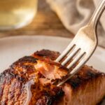 A close-up of a fork flaking a piece of cooked salmon, showing a juicy pink interior and a sticky, caramelized dark glaze on the outside.