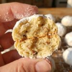 A close-up of a hand holding a powdered sugar snowball cookie broken in half, showing a buttery, nutty interior.
