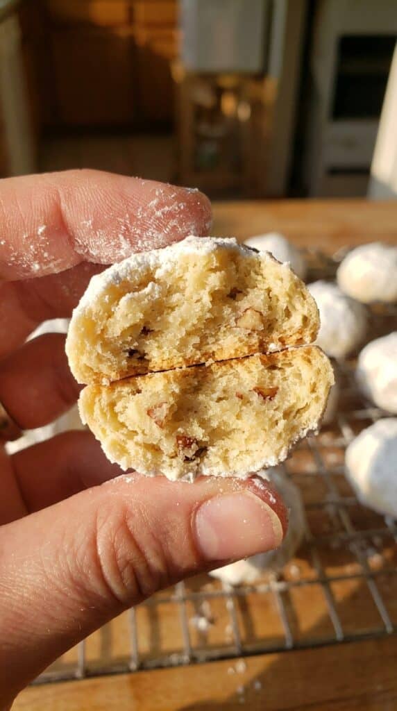 A close-up of a hand holding a powdered sugar snowball cookie broken in half, showing a buttery, nutty interior.