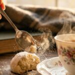 A close-up of a spoon breaking open a delicate, sugar-coated snowball cookie, showing its soft interior.