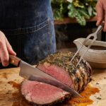 A close-up of a knife carving a thick slice of medium-rare prime rib, with savory juices pooling on a wooden cutting board.