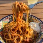 A close-up of a fork twirling cheesy taco spaghetti from a bowl, with a cheese pull and a dollop of sour cream.